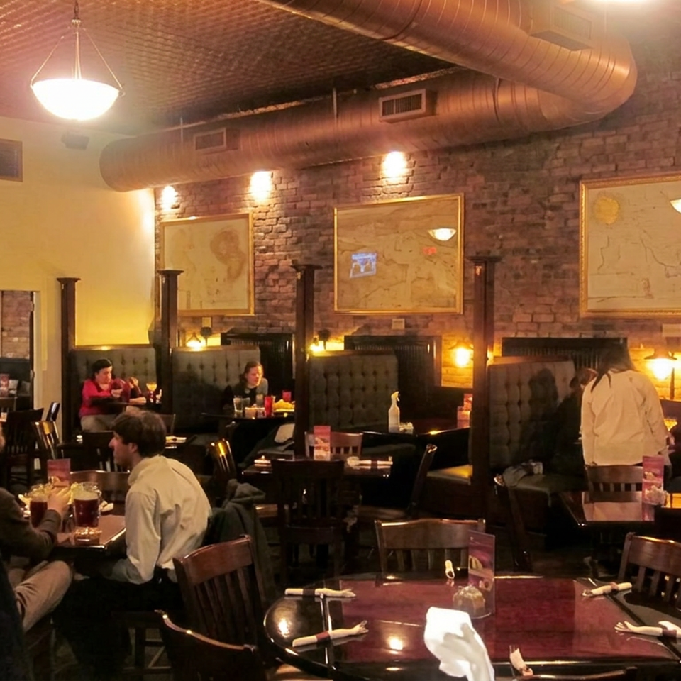 Warmly lit restaurant dining room with exposed brick walls, map artwork, booths, tables, and several diners.