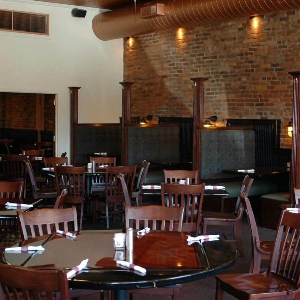 Spacious restaurant dining room with brick walls, exposed ductwork, wooden tables, upholstered booths, and neatly set place settings.