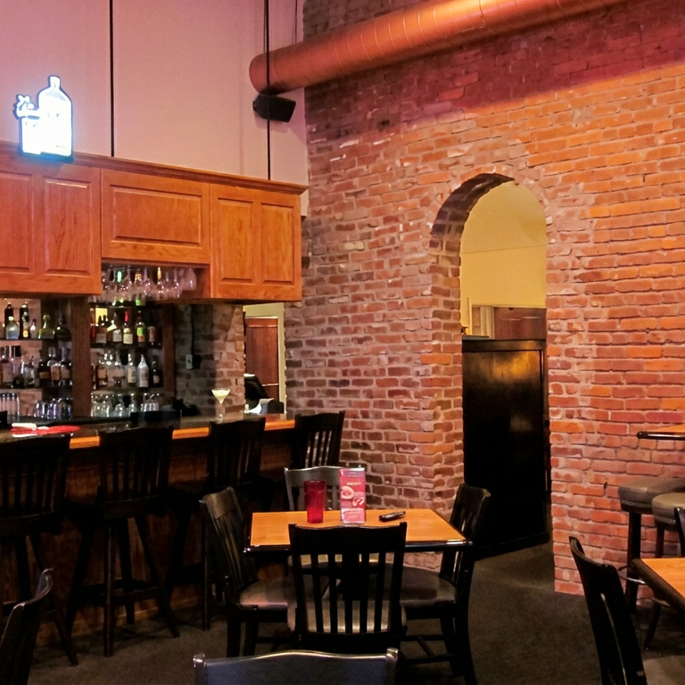 Warmly lit restaurant interior with exposed brick walls, wooden tables, black chairs, and a fully stocked bar.