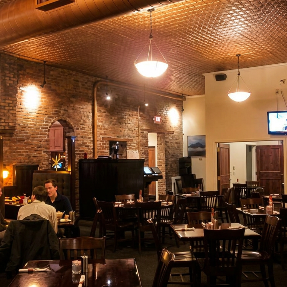 Warmly lit restaurant dining room with exposed brick walls, dark wooden tables, pendant lights, and diners seated in booths.